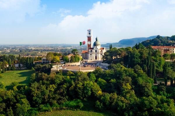 Un'antica torre con orologio, circondata da una vegetazione rigogliosa e uno sfondo urbano.