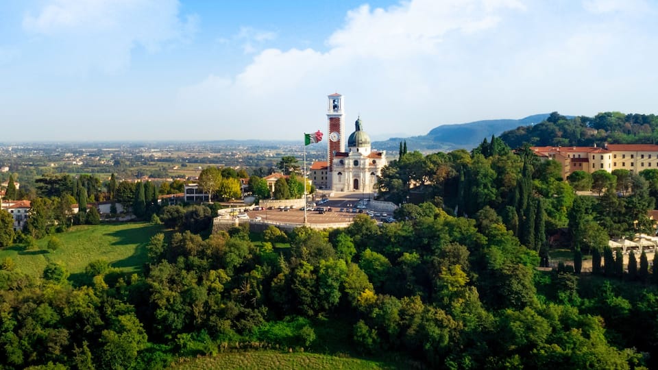 A historic tower with a clock, surrounded by lush greenery and a cityscape in the background.