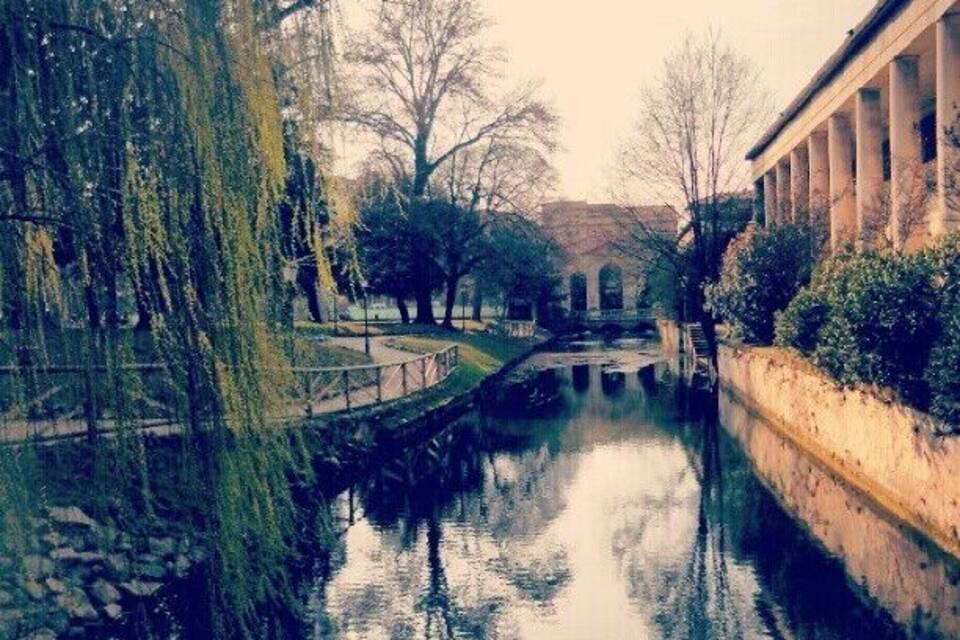 A calm canal with a stone bridge, willow trees, and a classical building with columns.