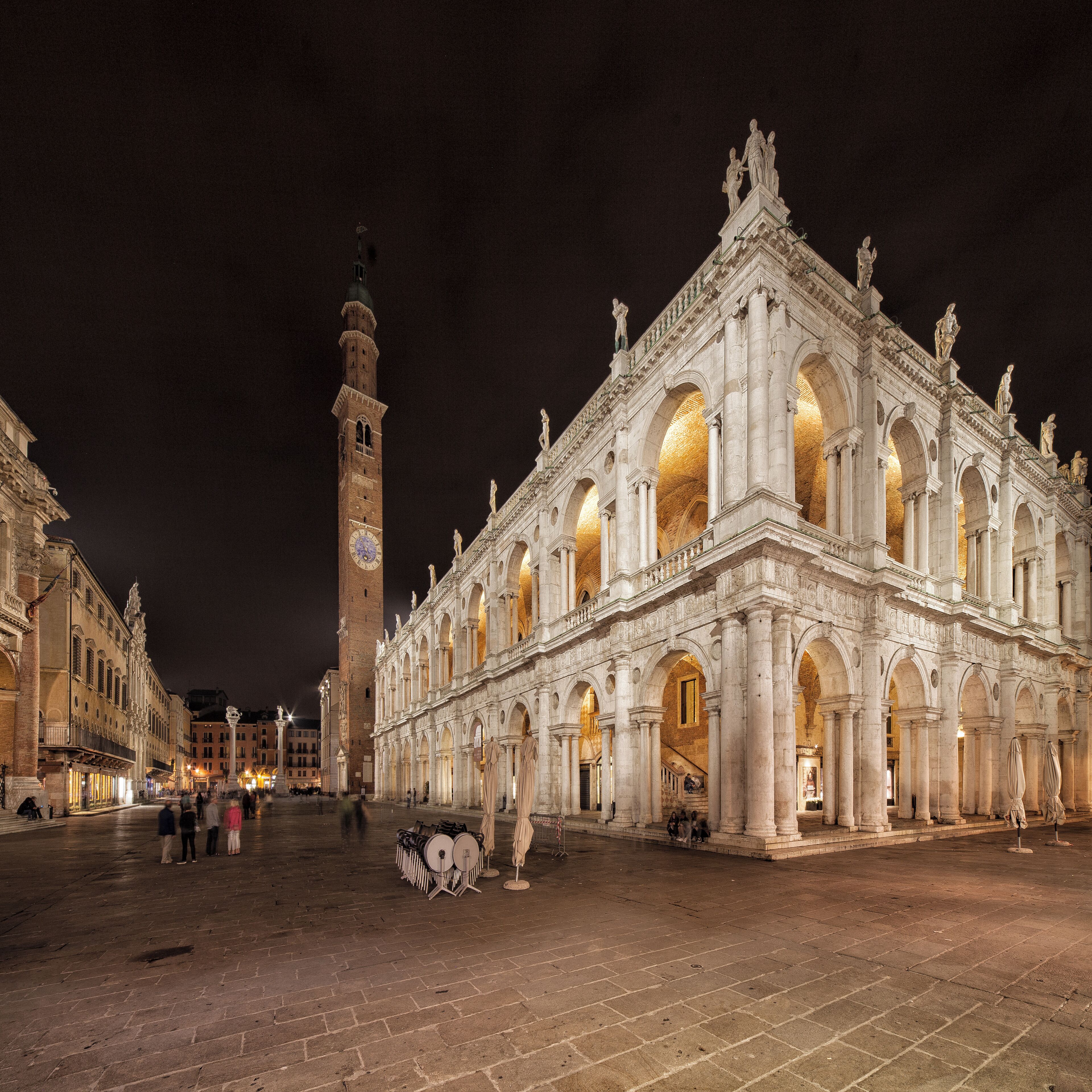 A historic building with arched windows and statues, a clock tower, and a square with people.