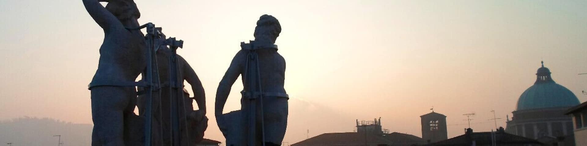 A statue of a man with a telescope and another statue of a man with a book, overlooking a cityscape with a prominent dome and buildings at sunset.