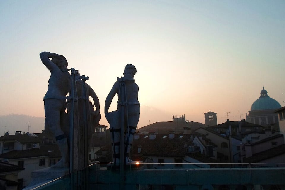 A statue of a man with a telescope and another statue of a man with a book, overlooking a cityscape with a prominent dome and buildings at sunset.