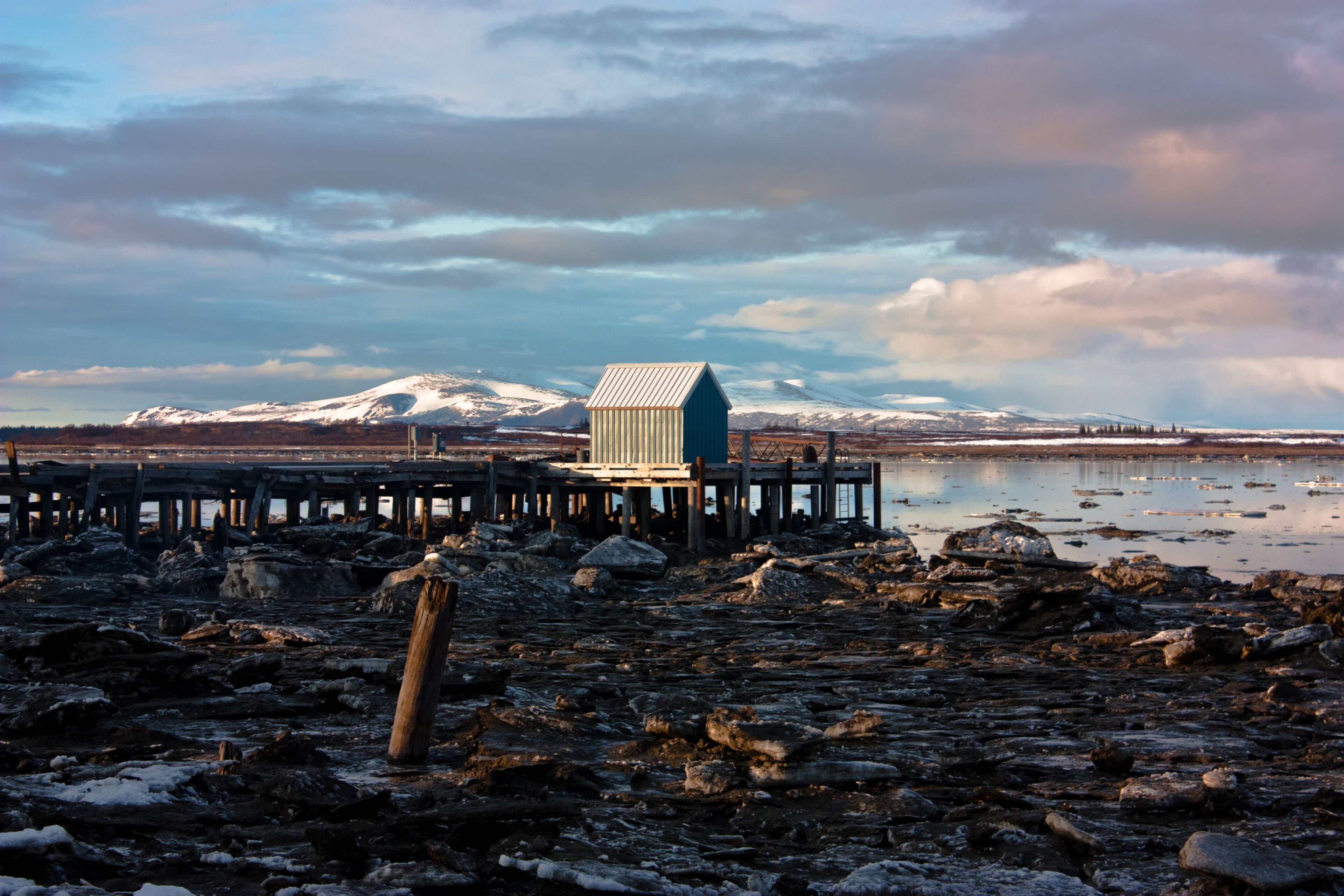 A wooden pier extending into a rocky, snow-covered shoreline with a small blue building at the end.