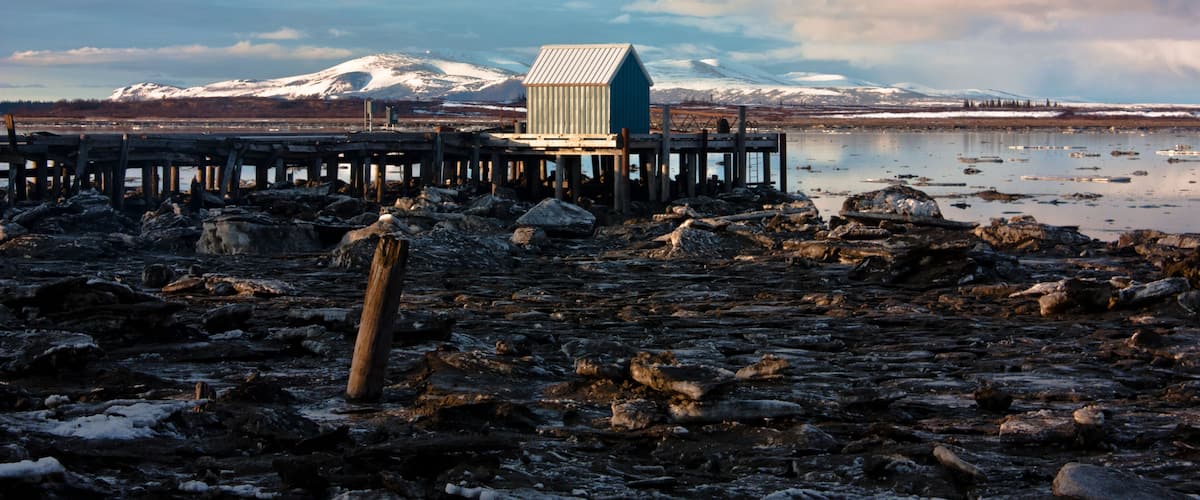 A wooden pier extending into a rocky, snow-covered shoreline with a small blue building at the end.