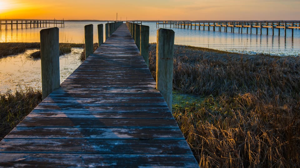 A wooden boardwalk leading to a pier extending into calm waters at sunset.