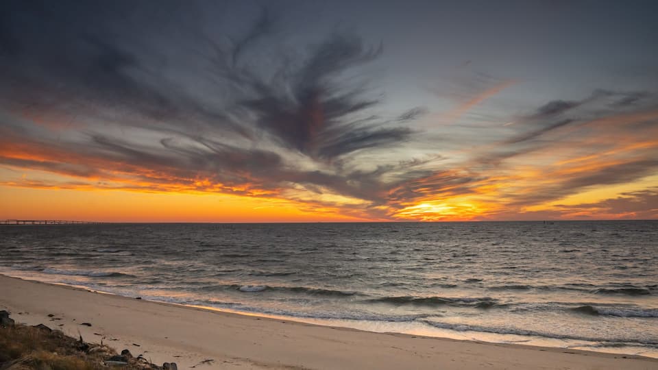 A sunset over a beach with waves, a pier in the distance, and a dramatic sky.