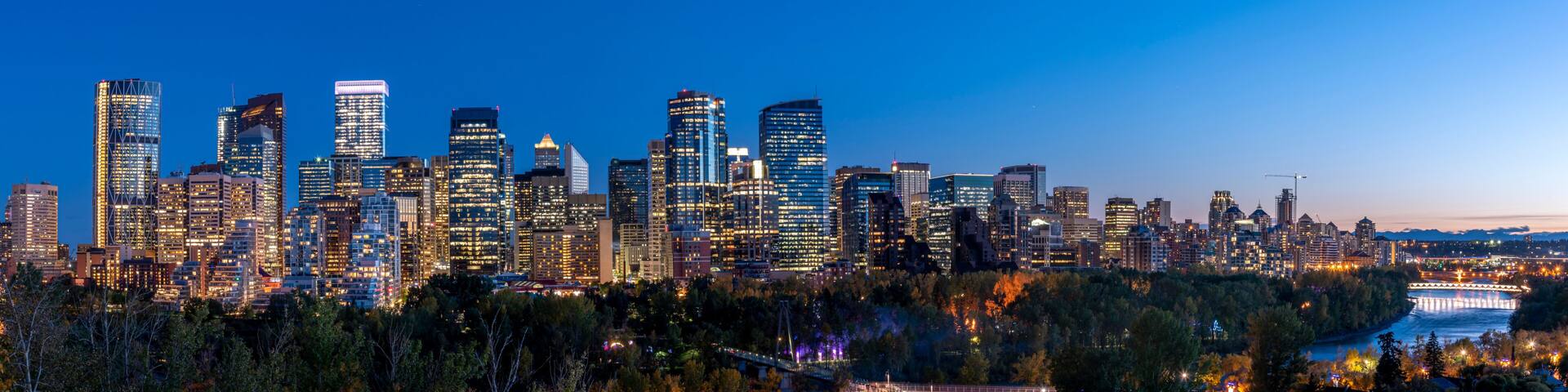 A city skyline at dusk with illuminated buildings and a river in the foreground.
