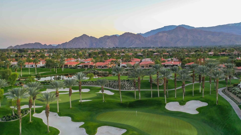 A golf course with sand bunkers, palm trees, and houses in the background.