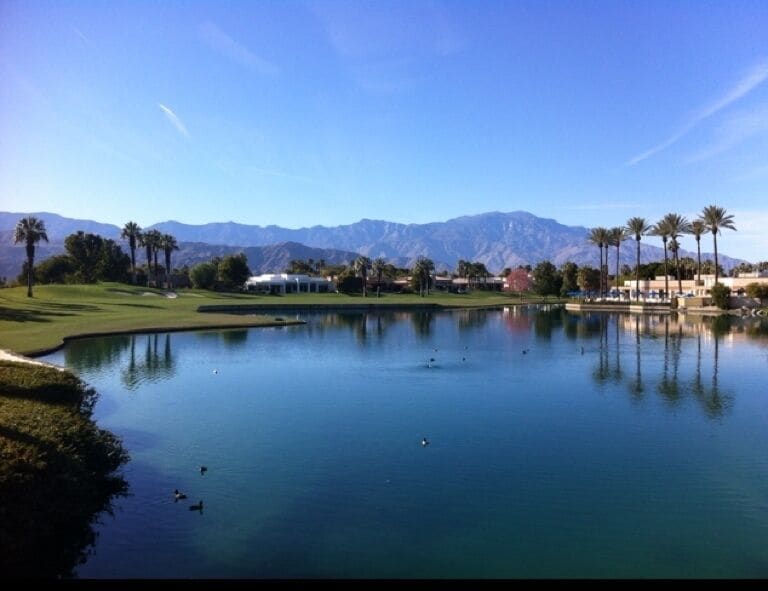 A golf course with a pond, palm trees, and mountains in the background.