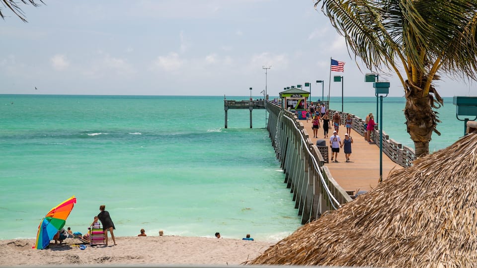 A beach with a wooden pier extending into the turquoise sea, palm trees, and people walking on the pier.