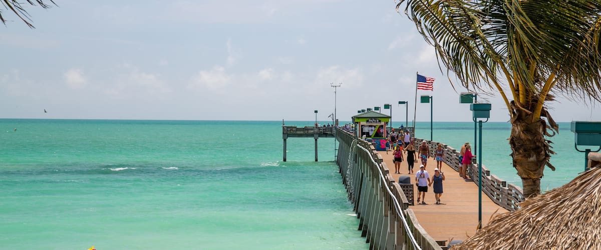 A beach with a wooden pier extending into the turquoise sea, palm trees, and people walking on the pier.