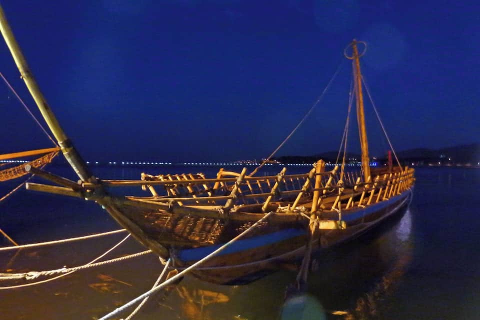 Una embarcación tradicional amarrada de noche, con el horizonte de una ciudad al fondo.