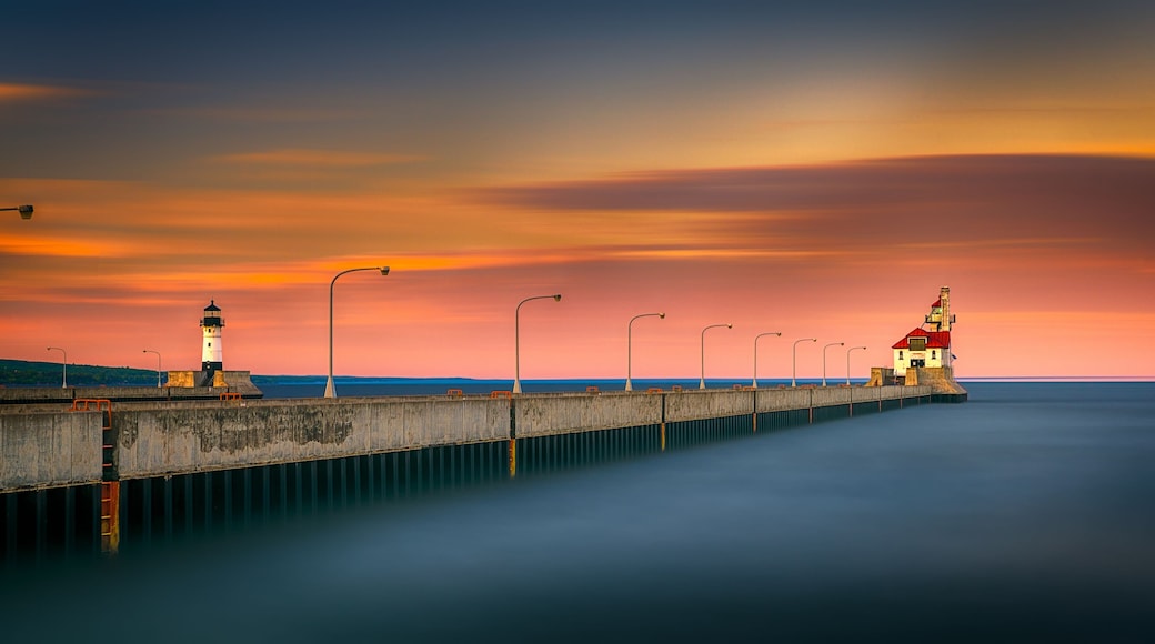 A pier with a lighthouse and a red-roofed building, extending into calm waters under a colorful sunset sky.
