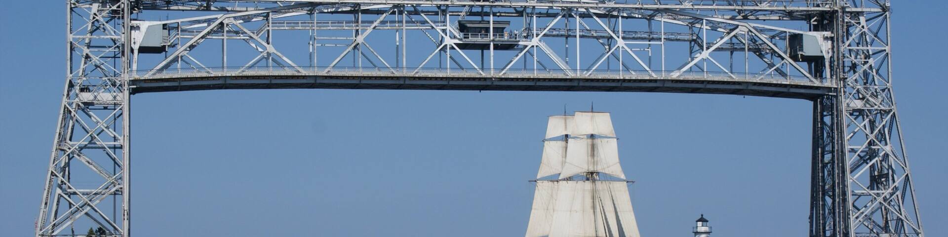 A large metal bridge spanning a body of water with sailboats and a lighthouse in the background.