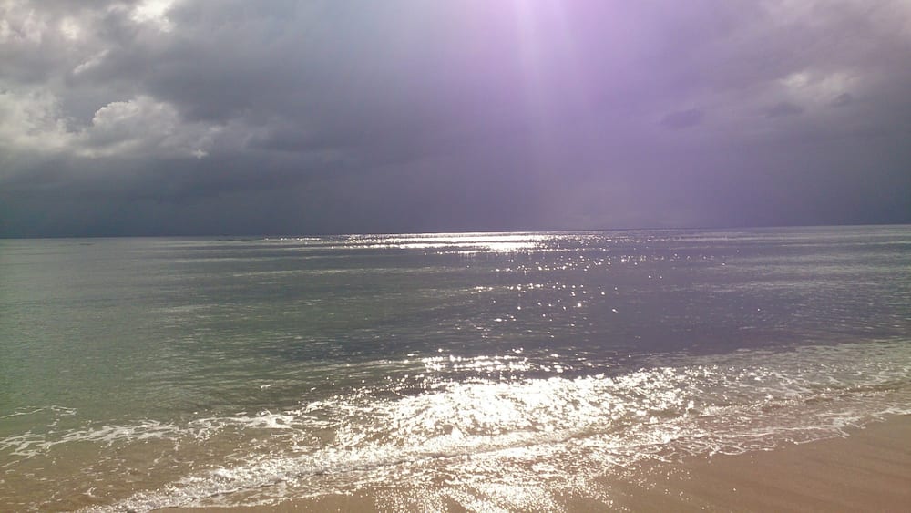 Une plage aux eaux calmes où se reflète la lumière du soleil, un ciel nuageux et un horizon lointain.