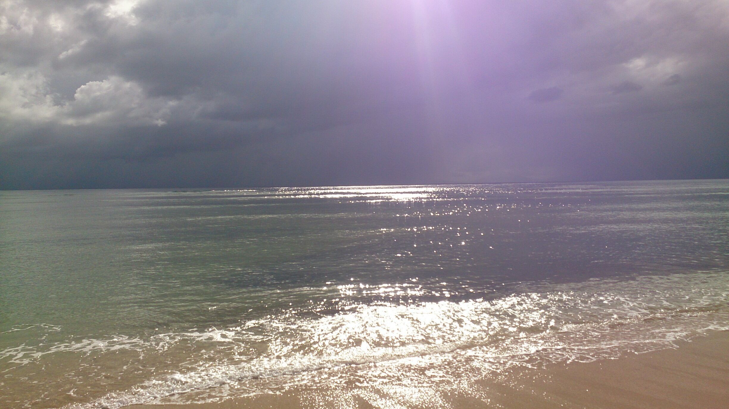 A beach with calm waters reflecting sunlight, a cloudy sky, and a distant horizon.