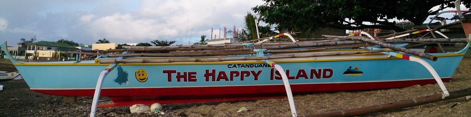 A colorful boat named "The Happy Island" with a smiling face and a palm tree on its side, docked on a beach with trees and buildings in the background.