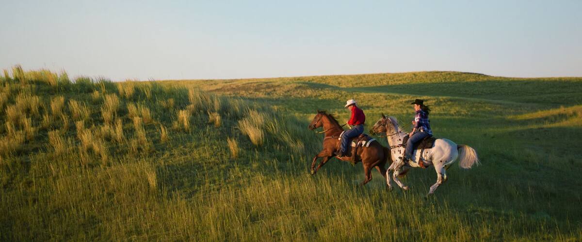 Two people riding horses across a grassy field with rolling hills in the background.
