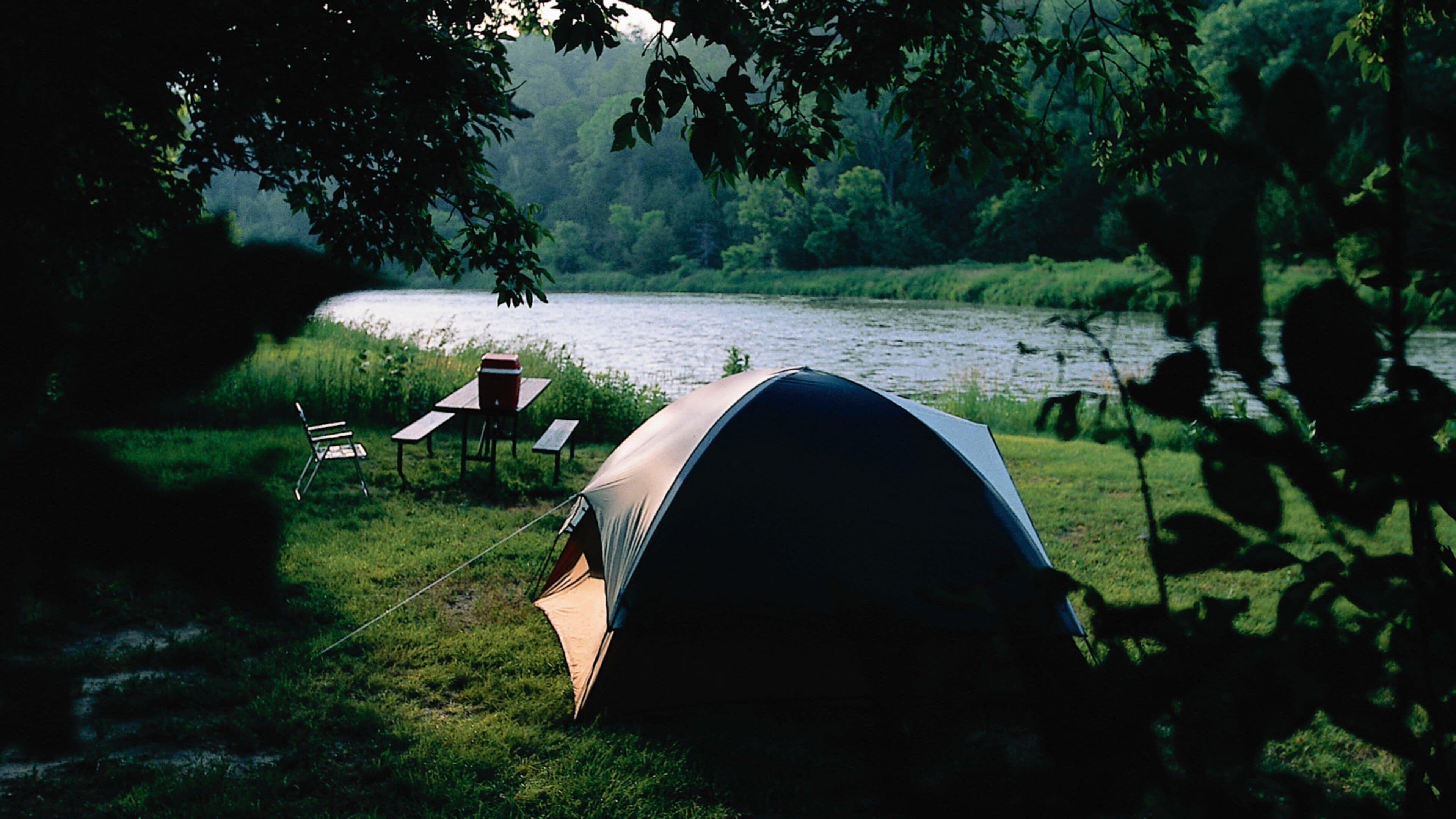 A tent set up by a lake with a table and chairs nearby.