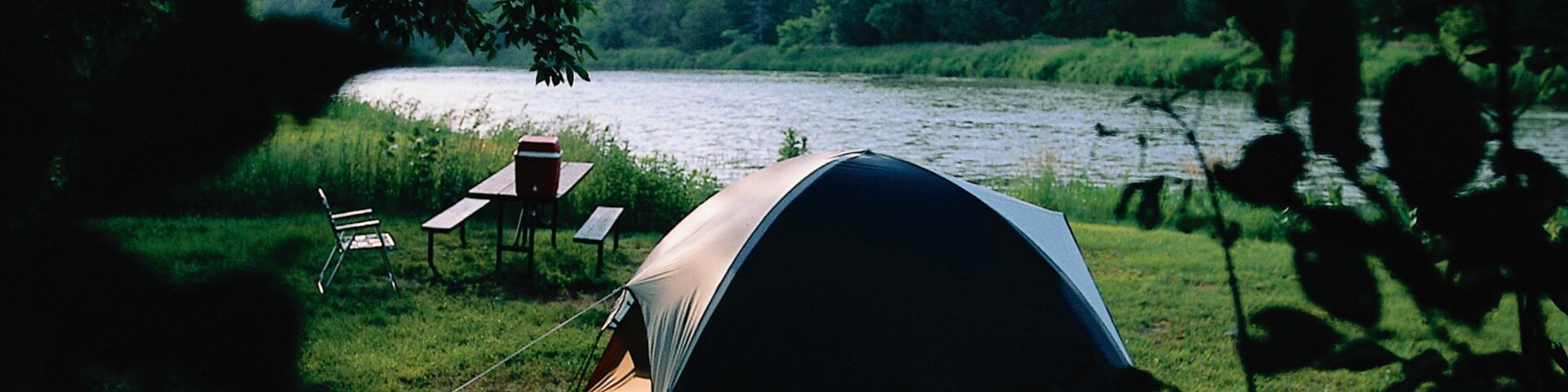 A tent set up by a lake with a table and chairs nearby.