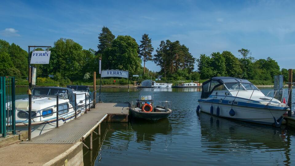 A marina with boats docked, a sign reading "FERRY," and a calm water body surrounded by trees.