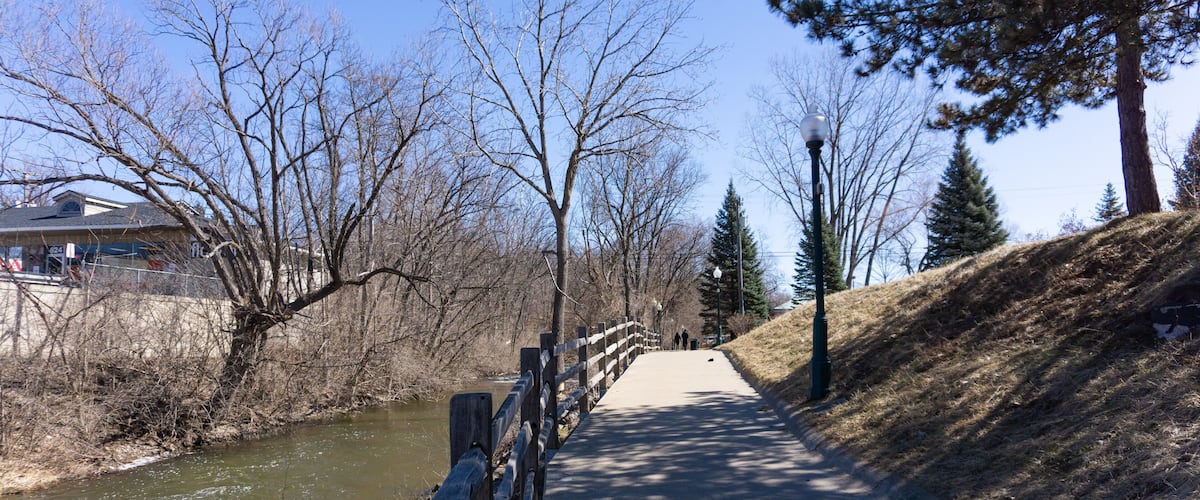 A riverside walkway with a wooden fence, trees, and a clear sky.