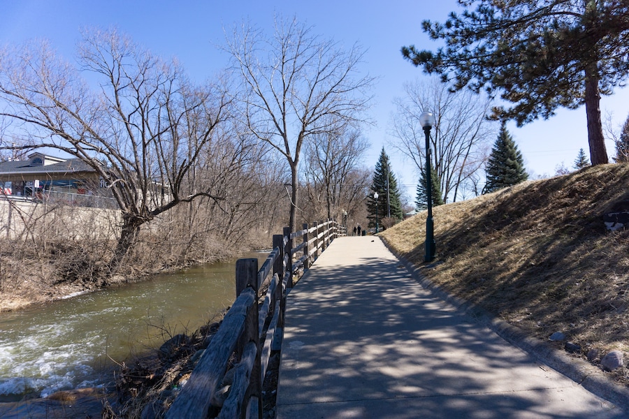 Un paseo junto al río con valla de madera, árboles y cielo despejado.