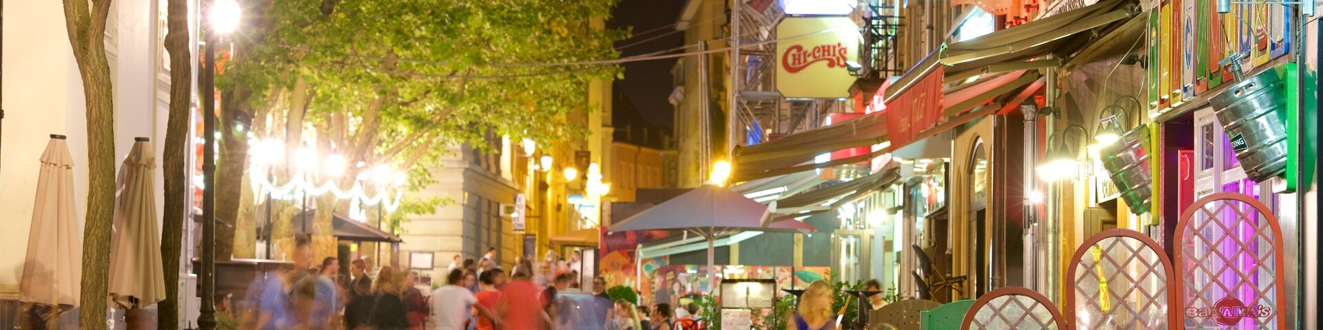 Uma rua movimentada à noite, com pessoas jantando em cafés ao ar livre, iluminada por postes e fachadas coloridas.