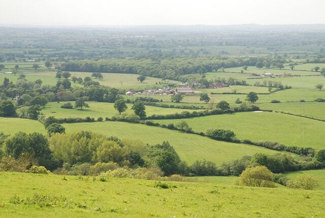 Rolling green hills, scattered trees, and a small village in the distance.