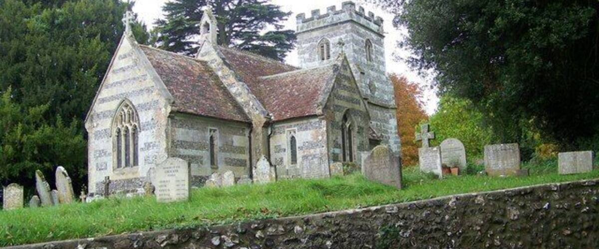 A stone church with a steep roof and a bell tower, surrounded by headstones and trees.
