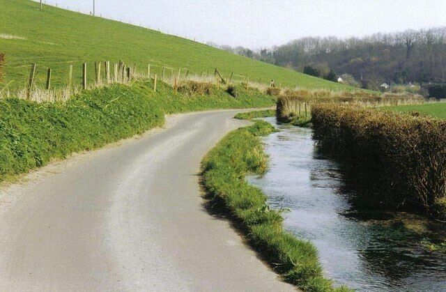 A narrow rural road curving alongside a small stream, bordered by grassy fields and a wooden fence.