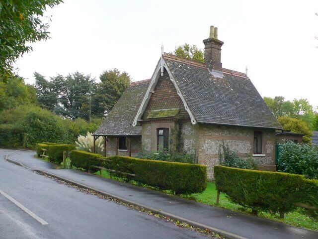 A quaint stone house with a steeply pitched roof, surrounded by well-maintained hedges and a road.