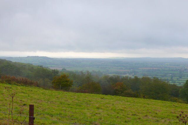 A grassy hillside with a fence, a distant view of rolling hills, and a cloudy sky.