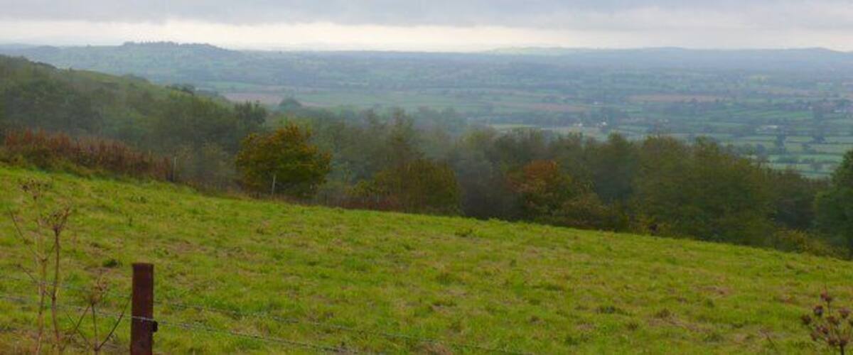 A grassy hillside with a fence, a distant view of rolling hills, and a cloudy sky.