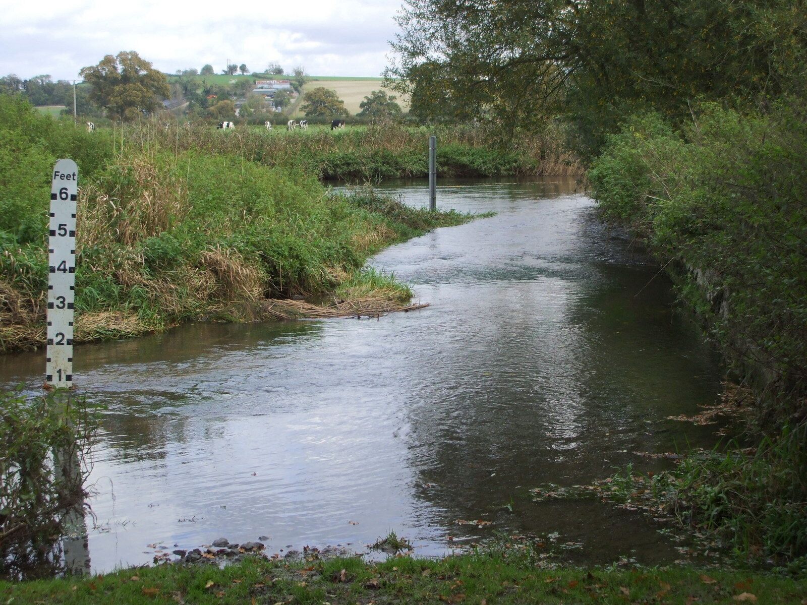 A narrow waterway with a footpath alongside, surrounded by greenery and a rural landscape in the background.