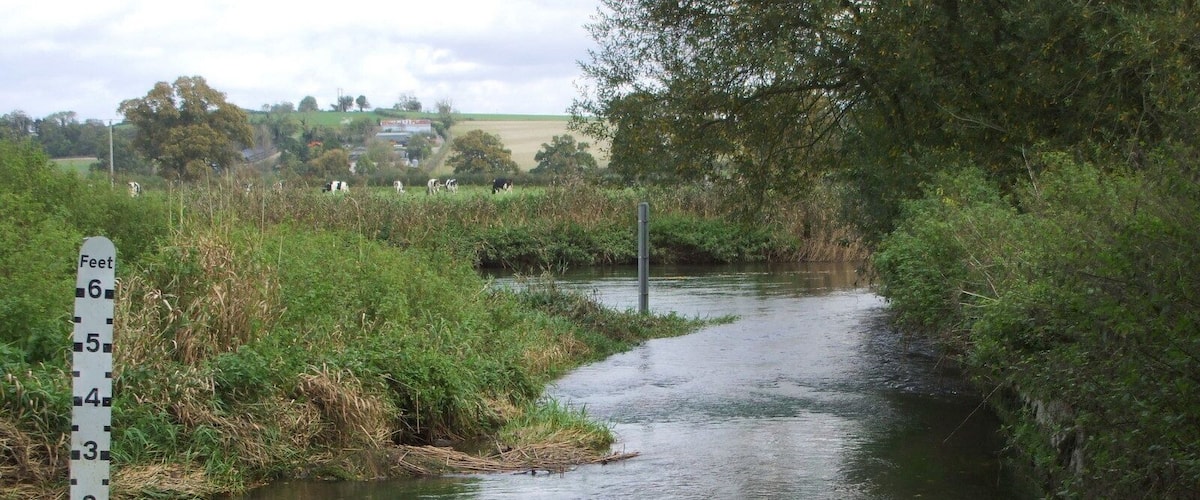 A narrow waterway with a footpath alongside, surrounded by greenery and a rural landscape in the background.