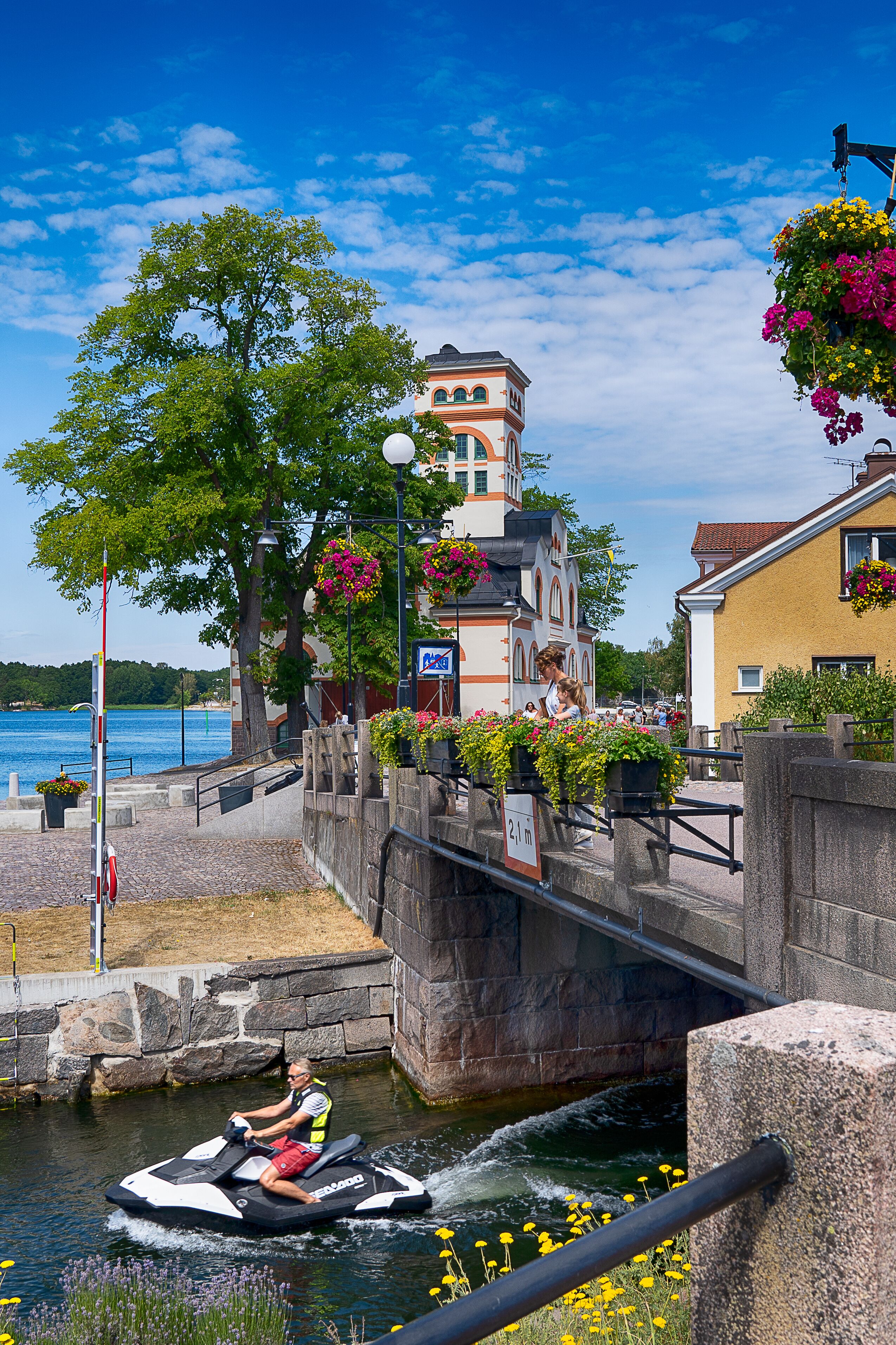 A canal with a boat, a bridge, and buildings with flower baskets.