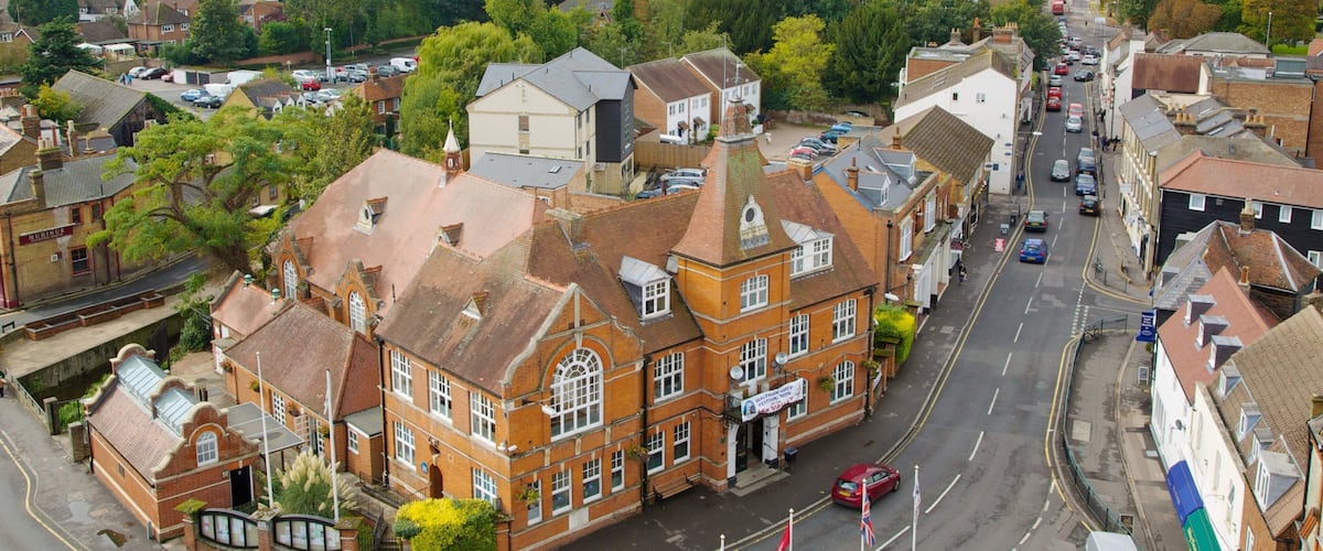 A town with a prominent red brick building, a main road, and surrounding greenery.