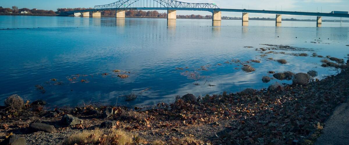 En stålbuebro over en rolig flod med efterårsblade ved bredden.