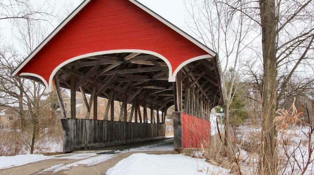 Snowy winter Wisconsin countryside landscape. Scenic view with old style bright red color covered bridge over stream during cloudy day. Waunakee, Madison area, Wisconsin, Midwest USA.