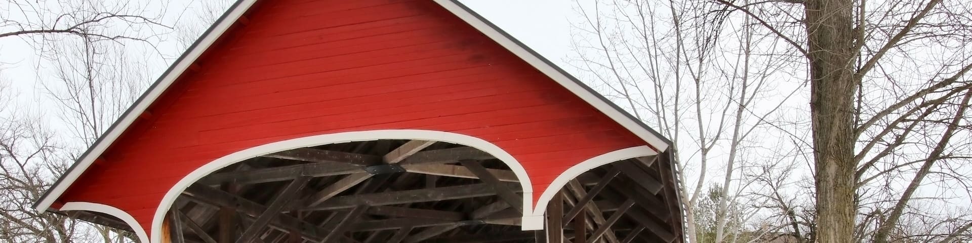 Snowy winter Wisconsin countryside landscape. Scenic view with old style bright red color covered bridge over stream during cloudy day. Waunakee, Madison area, Wisconsin, Midwest USA.