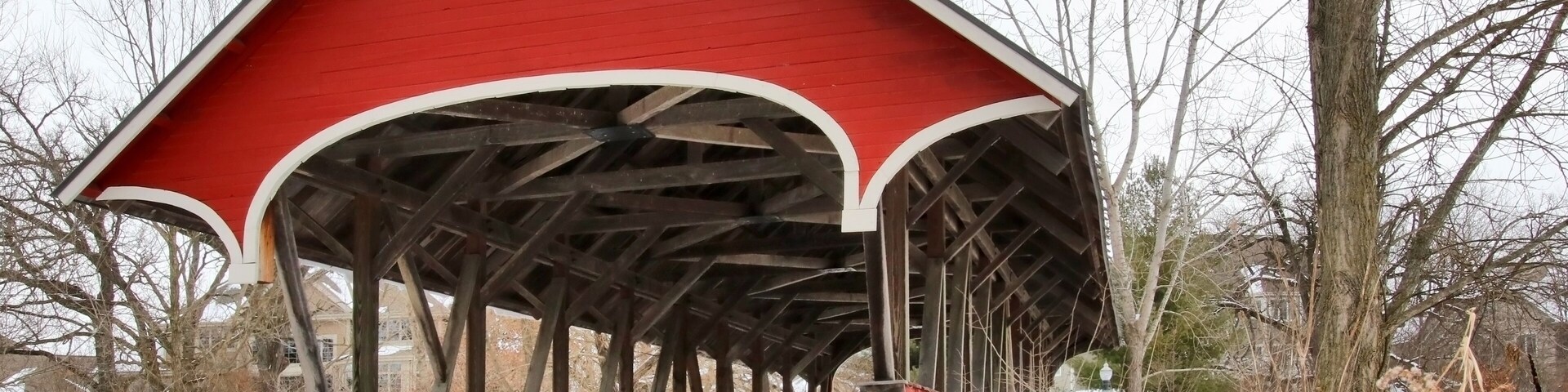Snowy winter Wisconsin countryside landscape. Scenic view with old style bright red color covered bridge over stream during cloudy day. Waunakee, Madison area, Wisconsin, Midwest USA.