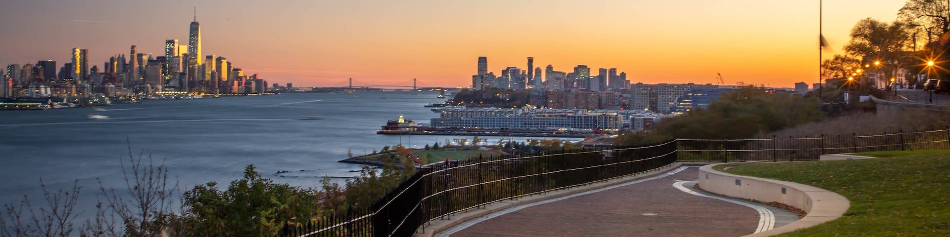 A sunset view of a city skyline across a river, with a park and a curved walkway in the foreground.
