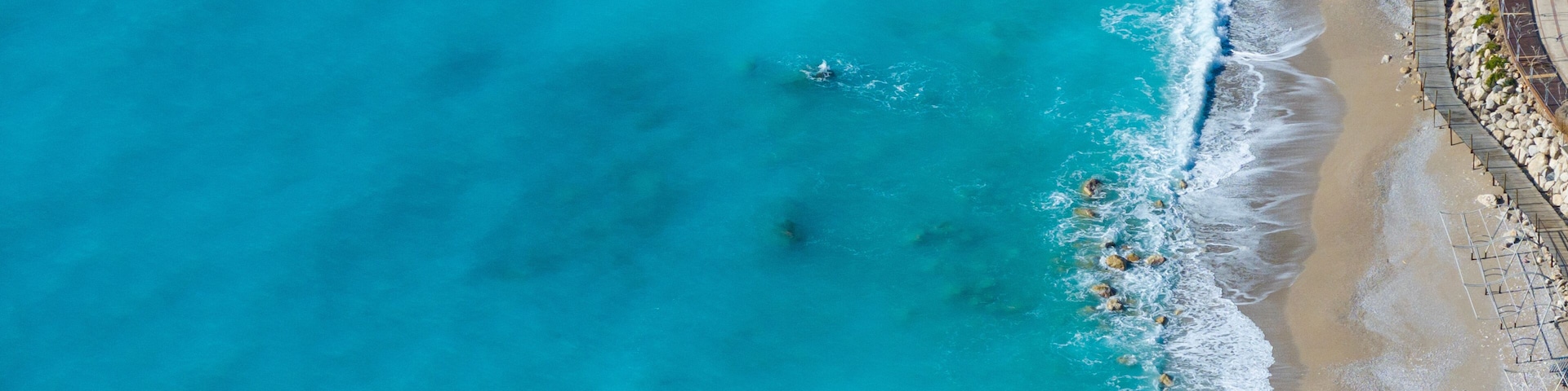 Una playa costera con aguas turquesas cristalinas, un rompeolas rocoso y un paseo marítimo pavimentado a lo largo de la orilla.