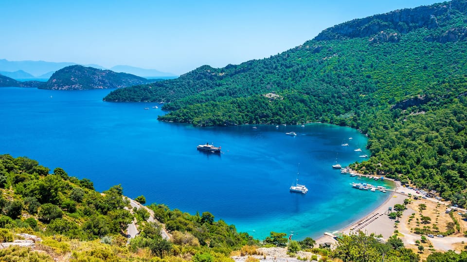 A coastal landscape with a clear blue sea, boats, and a forested island.