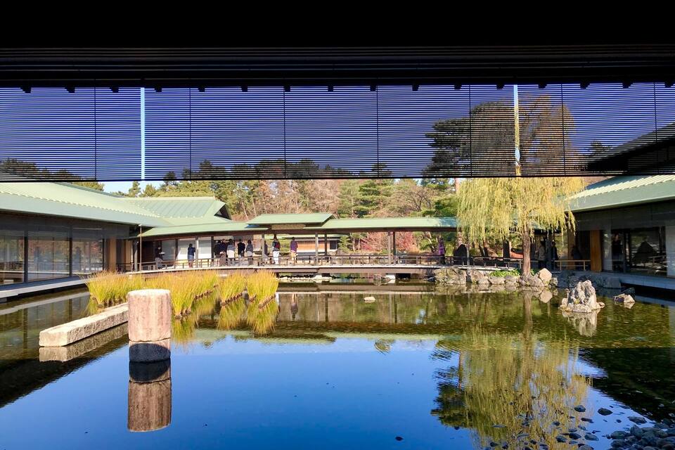 Een serene vijver met spiegelingen, omgeven door traditionele architectuur en een treurwilg.