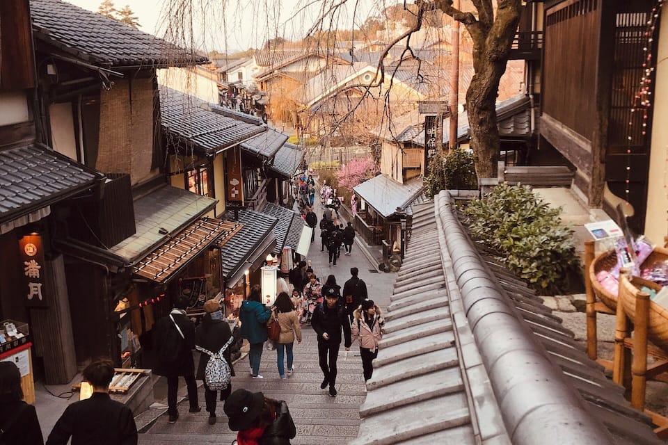 Een traditionele Japanse straat met voetgangers, houten gebouwen en een grote boom.