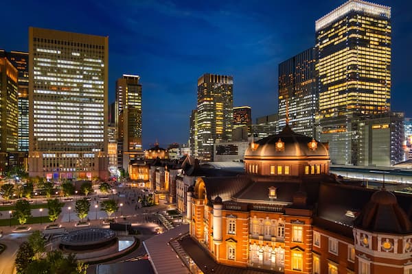 Une vue nocturne d’une ville, avec des gratte-ciel illuminés, un bâtiment historique surmonté d’un dôme et une place bien éclairée.