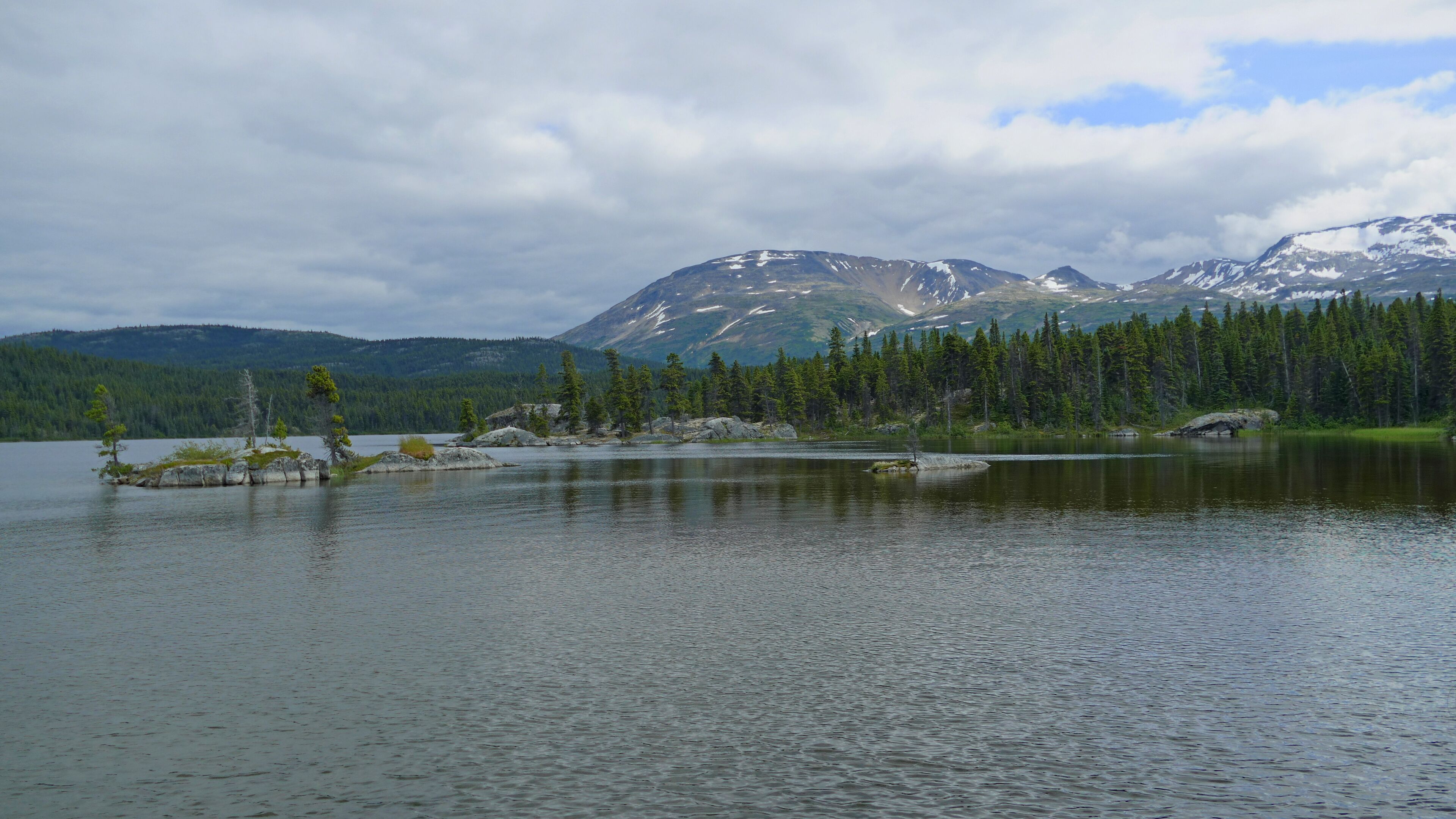 A calm lake surrounded by dense forests and snow-capped mountains under a cloudy sky.