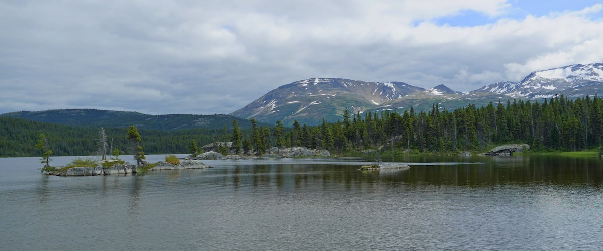 A calm lake surrounded by dense forests and snow-capped mountains under a cloudy sky.